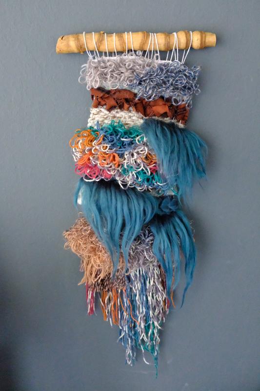 a woven wall hanging on a bamboo dowel rod with looped yarn in various greys, blues, greens, oranges, red and neutral browns too! gorgeous dark teal fiber inspired by waterfalls spill over the looped sections. 