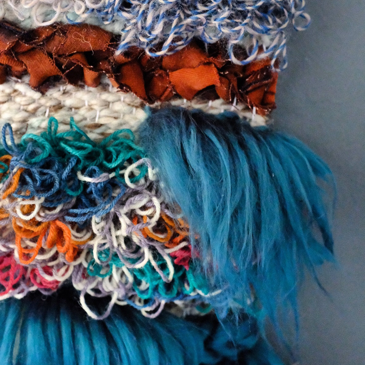 a close up of the dark teal fiber waterfall, looped yarn in various colors, and upcycled bleached demin in a burnt sienna color braided above the waterfall.  